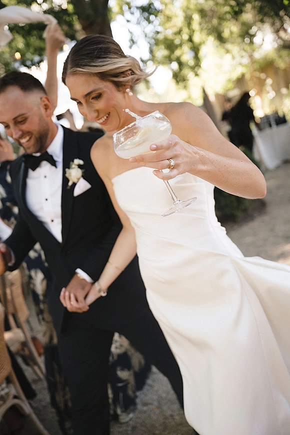 Wedding reception moment with bride holding champagne coupe, newlyweds laughing in tux and strapless dress under bistro lights among guests
