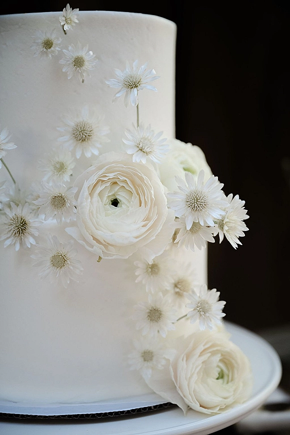 Wedding cake with white wedding cake buttercream frosting topped with white ranunculus and daisy flowers on a plate against a dark background
