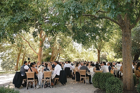 Outdoor wedding reception with long banquet tables in white linens and wicker chairs beneath trees, with string lights in sunlight