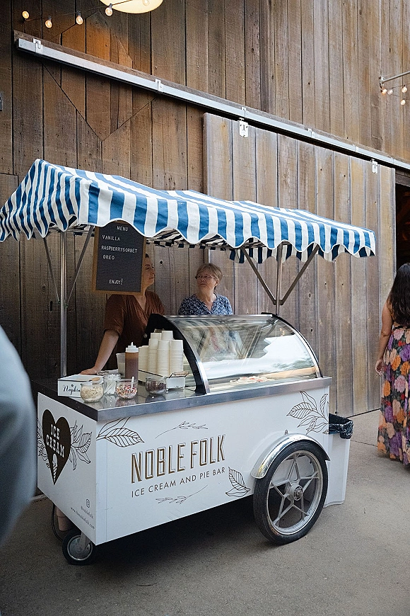 Ice cream cart wedding ice cream cart setup with striped canopy, menu sign, cups, and toppings jars against a barn wall with string lights