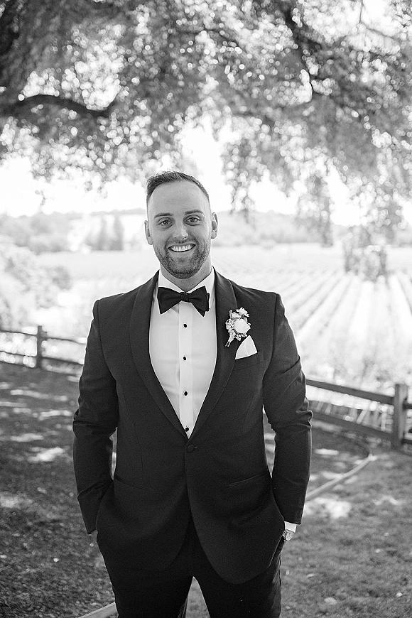 Groom portrait in a black tie tuxedo with bow tie and boutonniere, hands in pockets under a tree canopy by vineyard rows