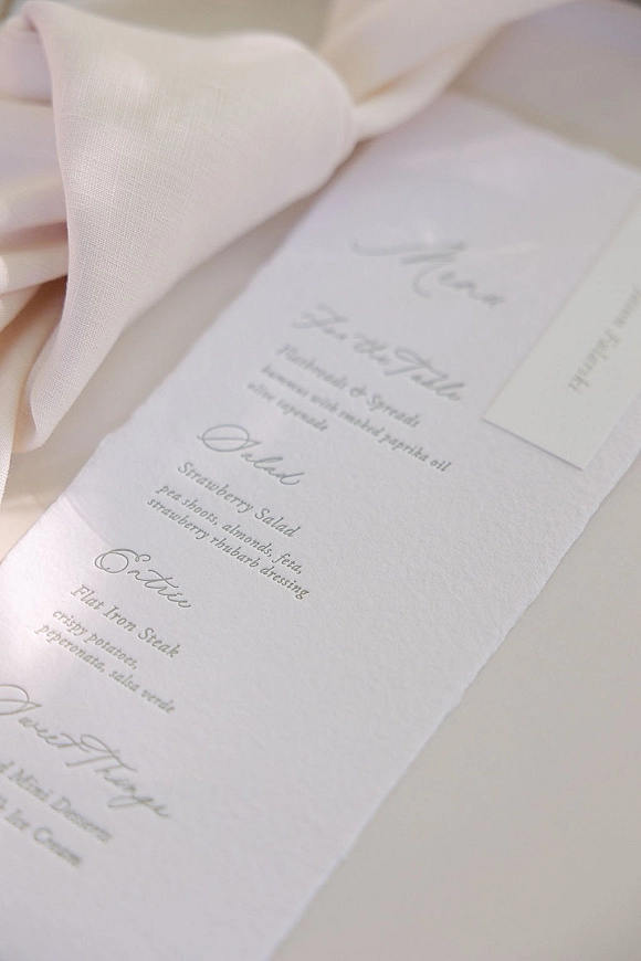 Wedding menu card with deckle edge paper and calligraphy text on a linen napkin atop a white tablecloth in a neutral flatlay