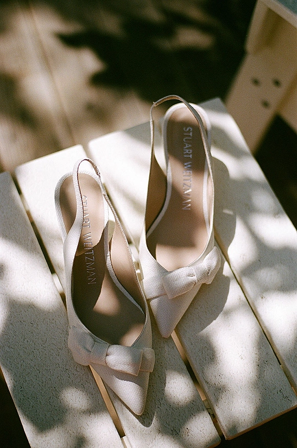 Bridal shoes, white wedding heels with bow detail and slingback straps on a white wooden surface with sunlit shadows in flat lay