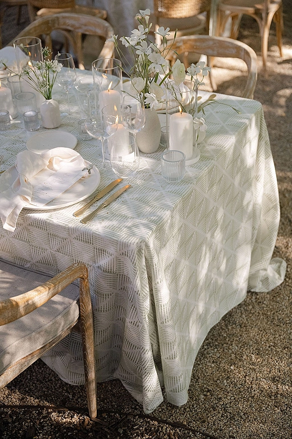 Reception tablescape with an outdoor wedding table setting, white plates and gold flatware, menu card, candles, and bud vases on gravel with rattan chairs