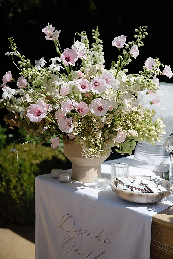 Wedding drink station with ribbed glass tumblers, votive candles, and a large floral arrangement on a white linen table in sunlit garden greenery
