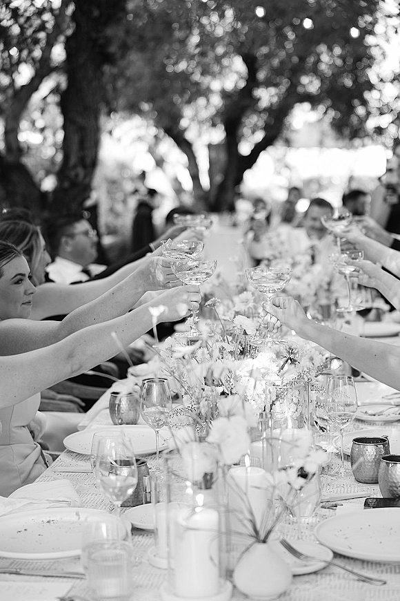 Wedding reception toast as guests raise champagne coupes over a long banquet table with floral centerpieces and candles beneath trees
