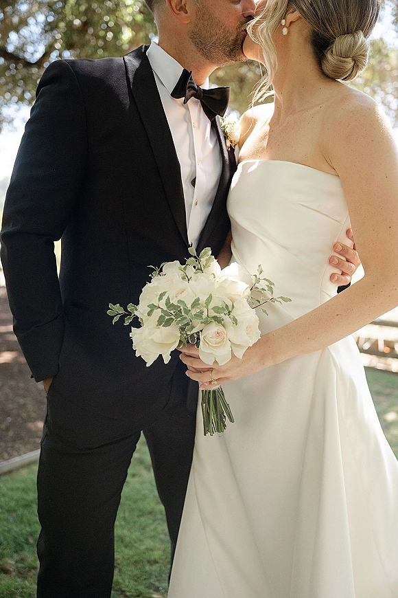 Wedding kiss portrait of bride and groom kissing, bride in strapless dress holding white rose bouquet, sunlit trees and fence behind
