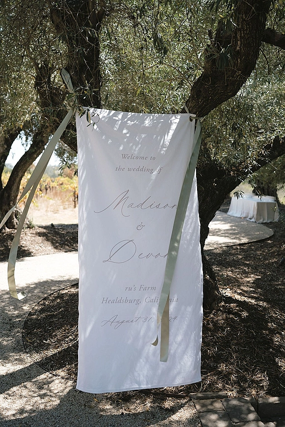 Wedding welcome sign, linen fabric banner with calligraphy lettering and ribbon ties hanging from tree branches along a garden pathway