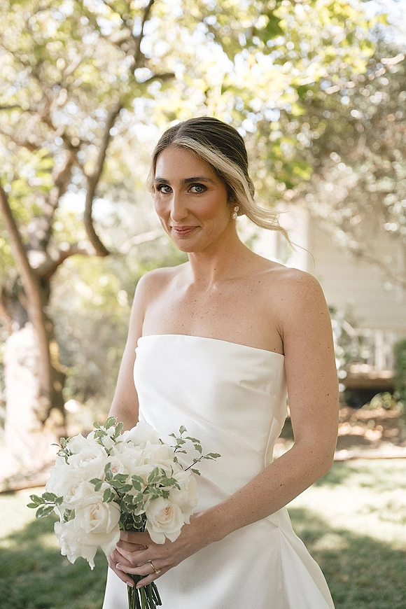 Bridal portrait of a bride holding bouquet of white roses and greenery in a strapless dress, pearl earrings, in dappled garden light