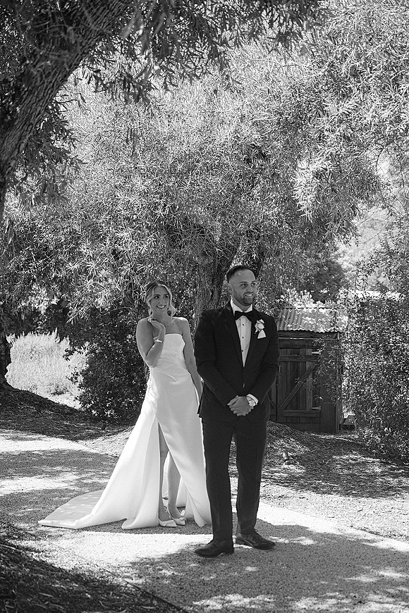 First look moment as bride taps groom’s shoulder on a sunny garden path under trees, her strapless dress and his tuxedo by a wooden gate