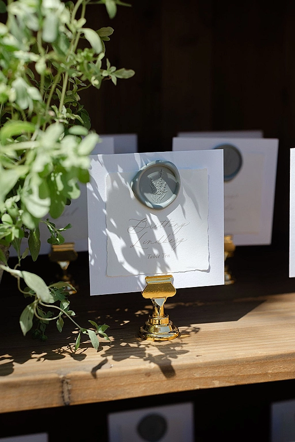 Wedding place card with calligraphy place cards styling, sage green wax seal on a gold holder with greenery against a dark backdrop