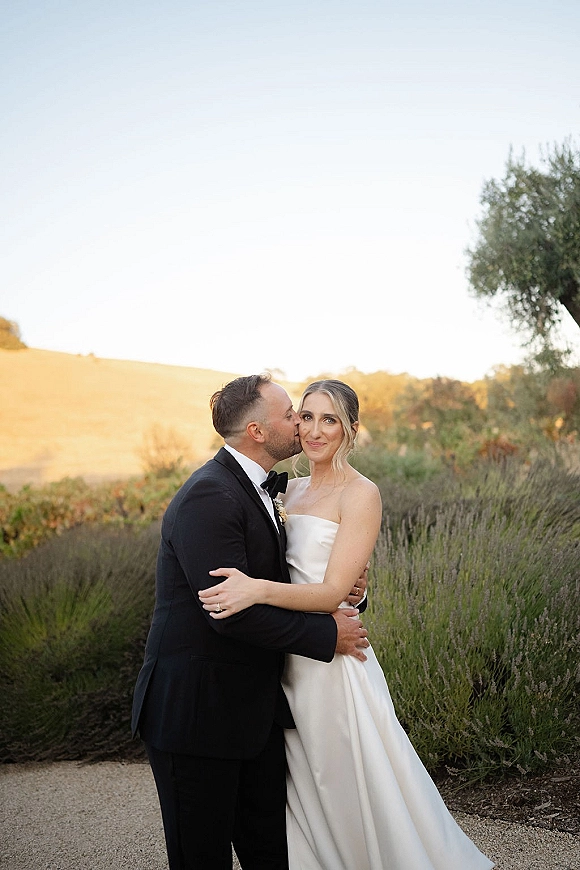 Couple portrait of groom kissing bride’s cheek as she smiles in a strapless wedding dress and black tuxedo on a hillside at sunset