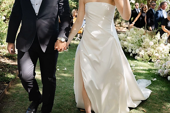 Wedding recessional as bride and groom holding hands walk down a flower-lined grass aisle with guests behind and rose petals scattered