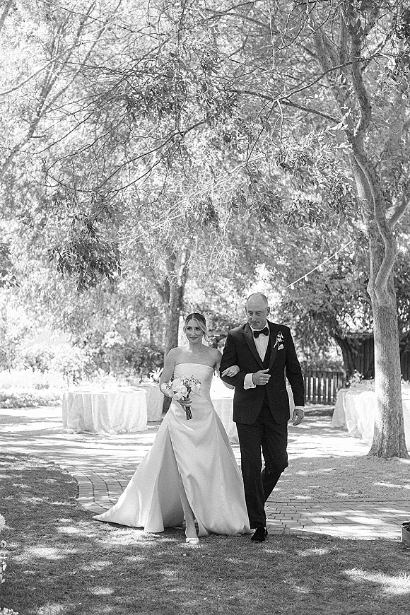 Wedding processional as bride walking down aisle with her father, holding a white bouquet, on a tree-lined brick walkway under string lights