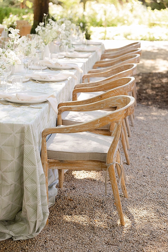 Reception tablescape with an outdoor wedding reception table featuring patterned cloth, white florals, candles, and wooden chairs in a garden