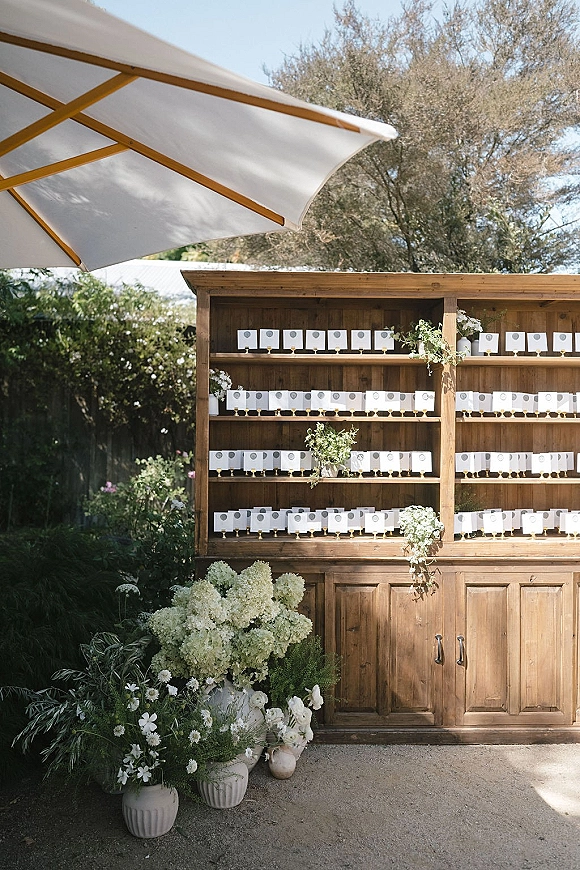 Escort card display with wedding escort cards on a wood shelving unit, framed by white floral arrangements and greenery on an outdoor patio under a large umbrella