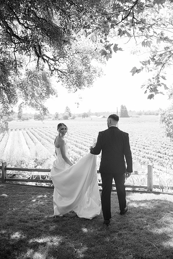 Wedding couple portrait in a black and white wedding photo, bride looking back as groom holds her dress train on a vineyard path by a wooden fence