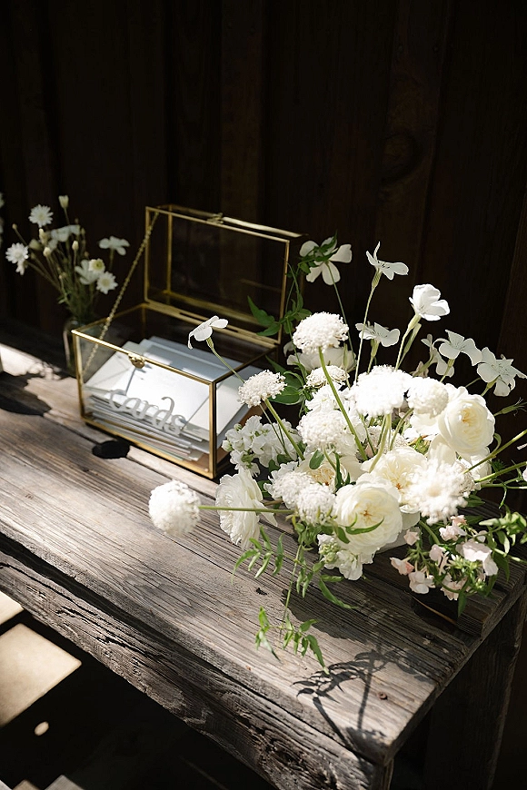 Wedding card box in gold glass with white flowers and greenery beside an acrylic sign on a rustic wooden table in sunlit shadows
