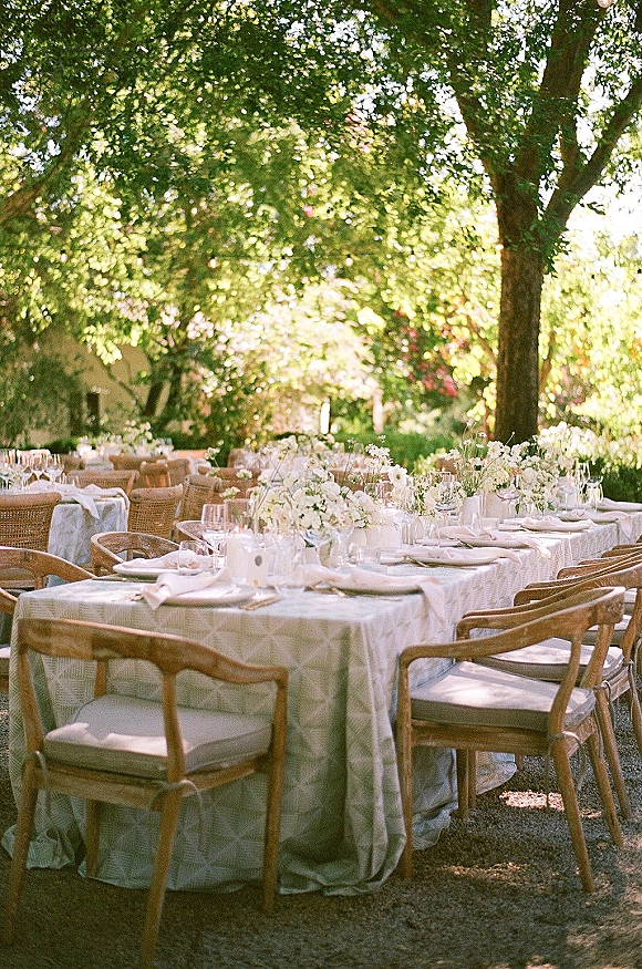Reception tablescape with white florals in bud vases, place cards, and glassware on linen tables under sunlit garden trees