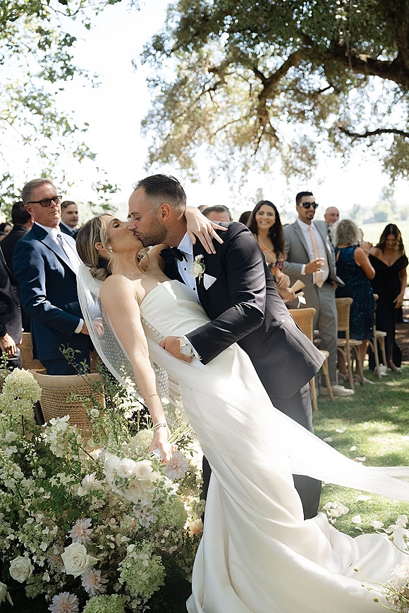 Wedding kiss as groom dips bride in a strapless dress and veil, bouquet of white roses and hydrangeas along outdoor aisle florals