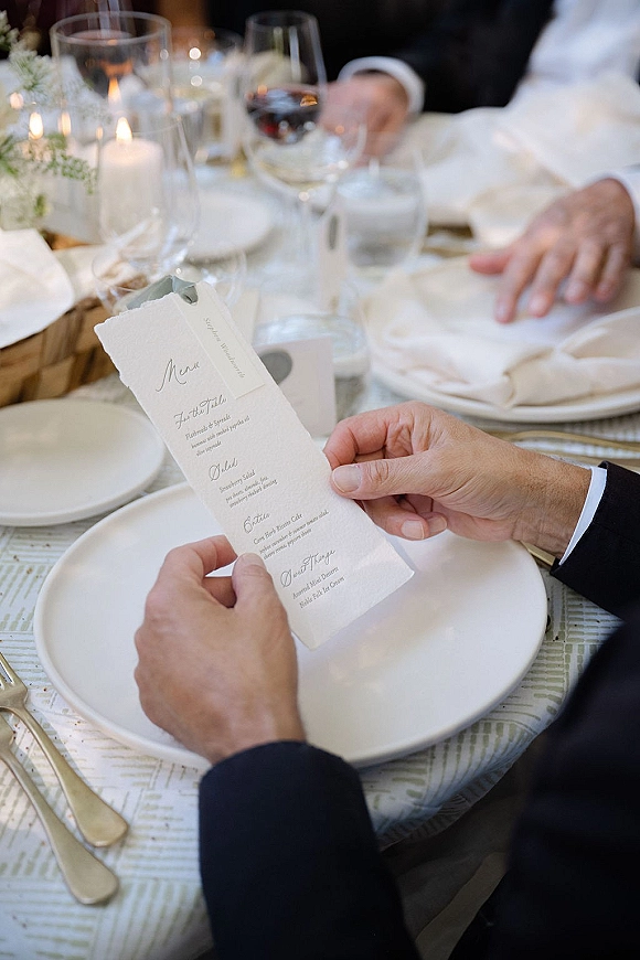 Wedding menu card with deckle edge paper at a reception place setting beside white plates, wine glasses, candles, and florals
