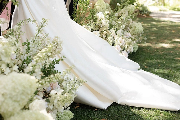 Wedding dress train draped over grass with bridal gown train detail beside white hydrangeas, blush blooms, and greenery in sunlight
