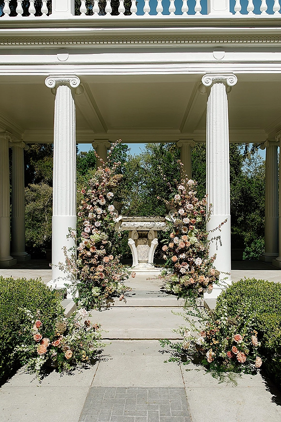 Ceremony floral arch with asymmetrical wedding arch flowers, roses and dahlias on floral pillars framing white portico steps, lush greenery.