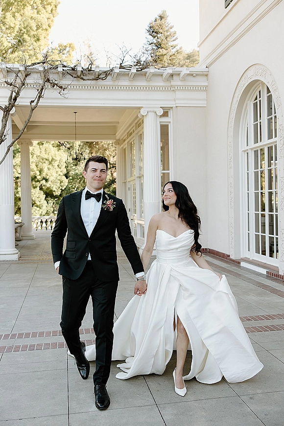 Couple portrait of bride and groom walking hand in hand, her strapless satin dress with a high slit, on a veranda with white columns
