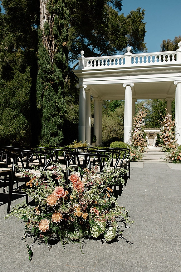 Outdoor ceremony setup with garden wedding ceremony aisle flowers, black chairs lining a stone aisle to a white column pavilion under blue sky