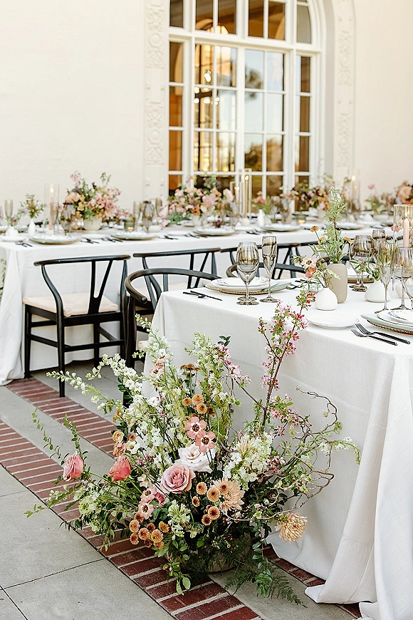 Reception tablescape with wedding head table decor featuring white linens, wildflower bud vases, pillar candles and black chairs on a stone patio