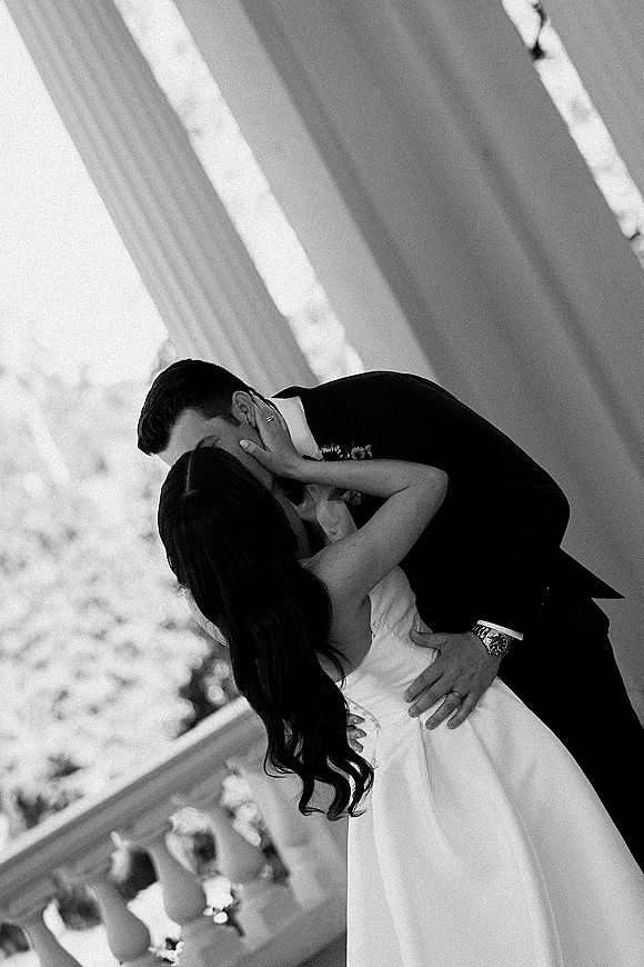 Wedding kiss as groom dips bride in a strapless dress, his boutonniere and watch visible on a columned porch with trees behind