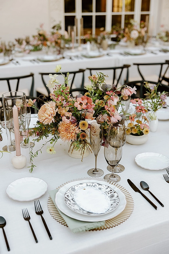 Reception tablescape with wildflower wedding centerpiece in bud vases, tapered candles, sage napkins, and patterned plates on a long table by windows