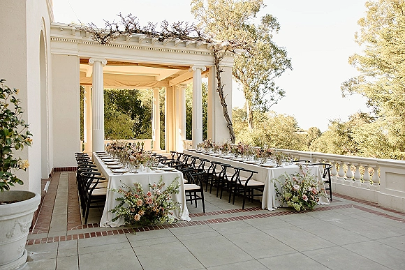 Reception tablescape on long banquet table wedding with white linens, black chairs, and low floral centerpieces under a covered terrace with columns