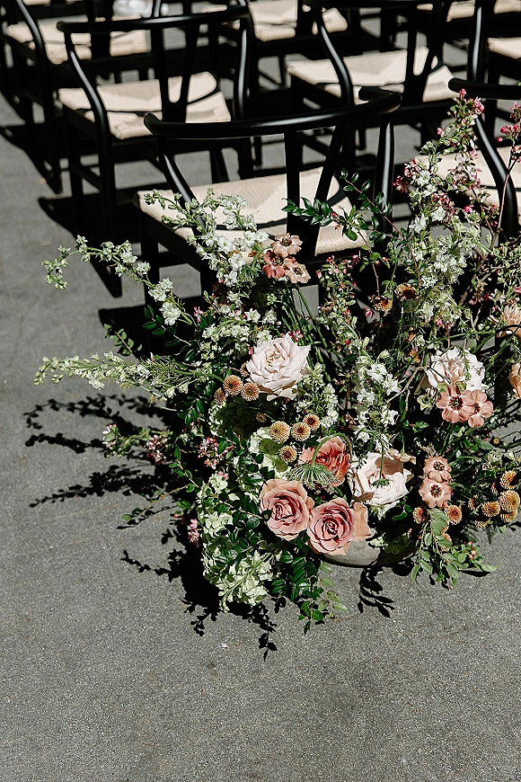 Ceremony aisle flowers with roses, greenery, and flowering branches arranged on the ground beside a paved aisle lined with black chairs