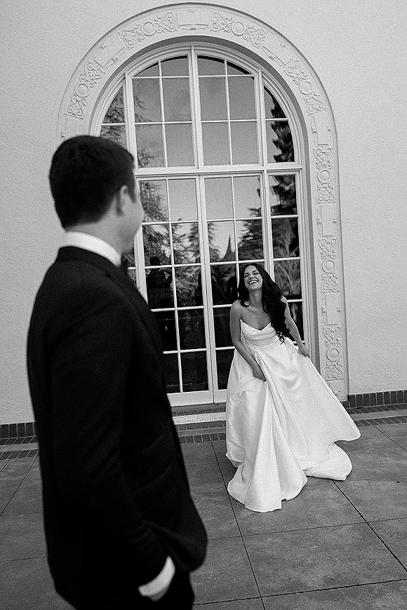 Couple portrait in black and white wedding portrait style, bride laughing in strapless gown as groom in tux looks at her by arched window patio