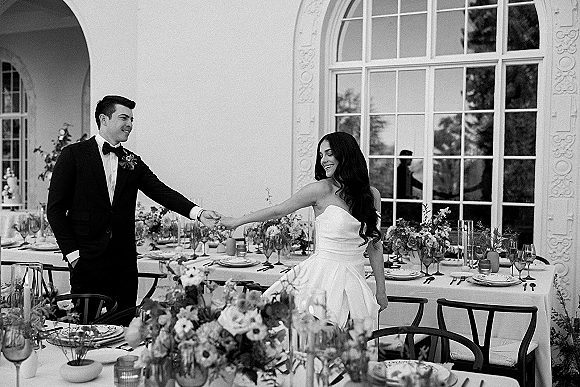 Couple portrait of bride and groom holding hands beside a long reception table with taper candles and floral centerpieces on an outdoor patio