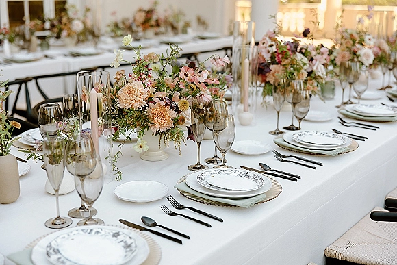 Reception tablescape with white linen wedding table setting, floral centerpieces, taper candles in glass holders, and layered place settings indoors