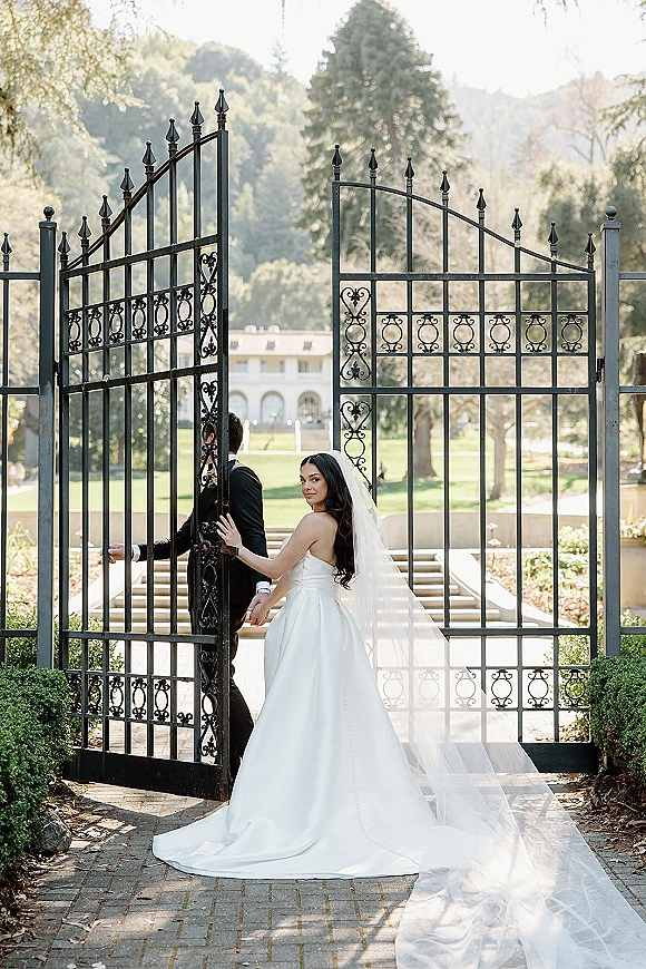Couple portrait of bride looking back as she and groom hold hands by a wrought iron gate, her cathedral veil trailing on garden path