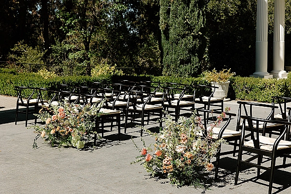 Ceremony seating with black chairs and white cushions lined in rows, flanked by aisle florals in an outdoor courtyard with hedges and columns