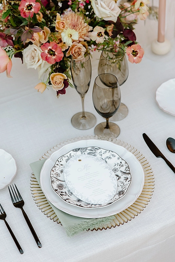 Wedding tablescape with a wedding place setting featuring floral centerpiece, smoked glass stemware, taper candle, and menu on white linen