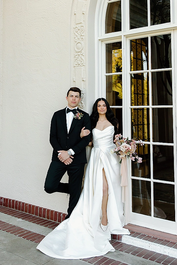 Couple portrait of bride and groom arm in arm, bride holding bouquet with trailing ribbon, beside an arched window and doorway