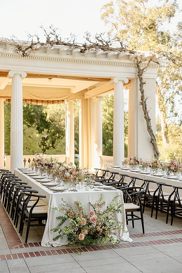 Reception tablescape with long banquet table in white linens, garden rose bud vases and greenery on a sunlit veranda with columns