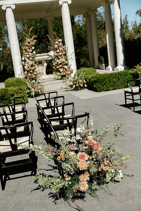 Ceremony setup with outdoor ceremony seating beneath a floral ceremony arch, black chairs lining a stone patio by a fountain pavilion