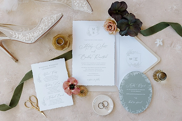 Invitation suite with deckle edge invitations arranged on neutral linen with sage green ribbon, wax seal, rings, perfume, and bridal shoes