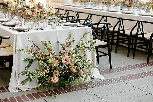 Reception tablescape with white linen, bud vase wedding centerpiece and greenery garland, layered place settings, and black chairs on a patio banquet table
