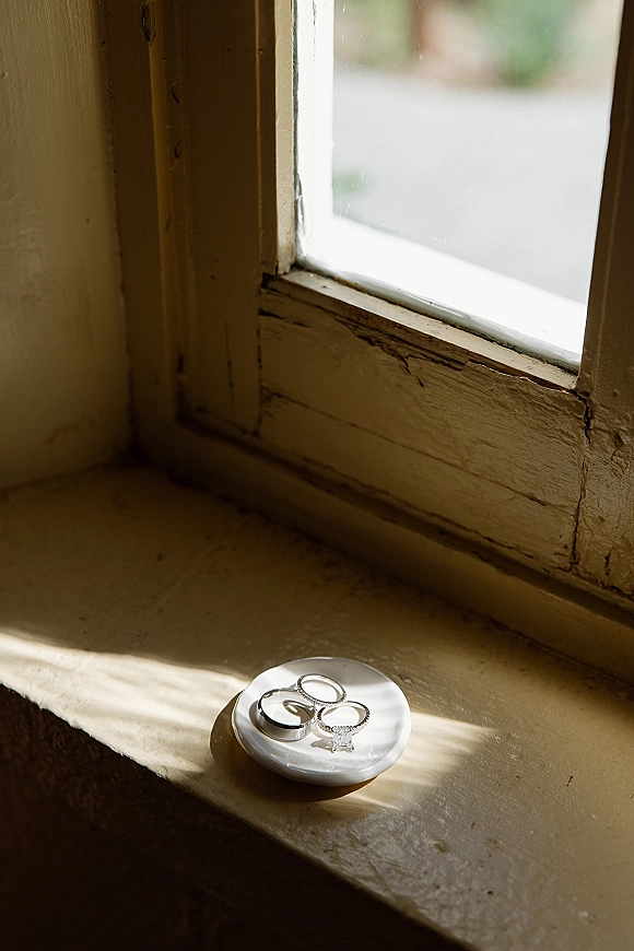 Wedding rings with an engagement ring close up stacked on a white ring dish on a windowsill in soft natural window light