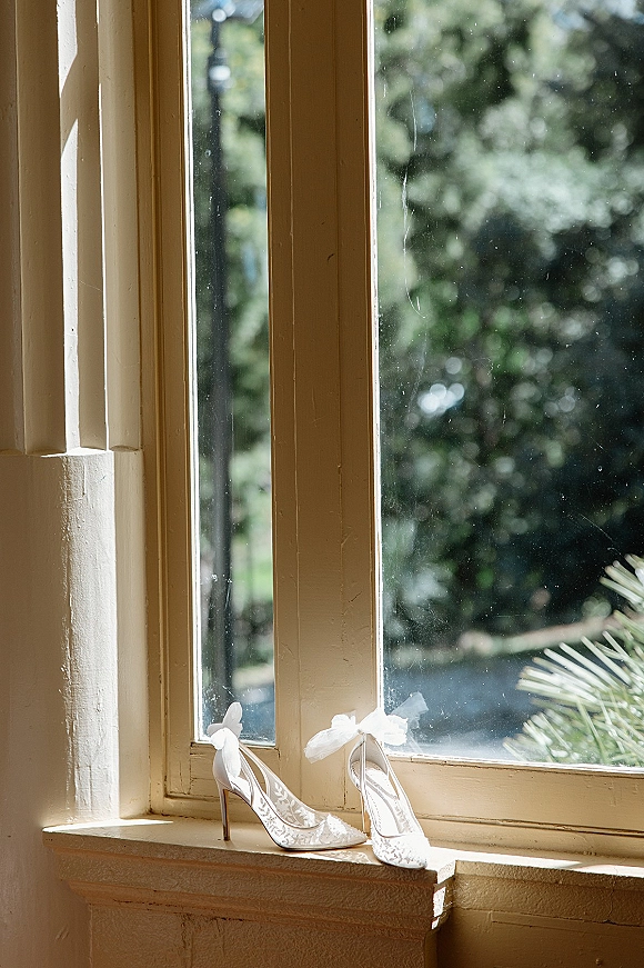 Wedding shoes with sheer bow ankle ribbons and embroidered mesh resting on a sunny windowsill beside a wooden frame and garden greenery