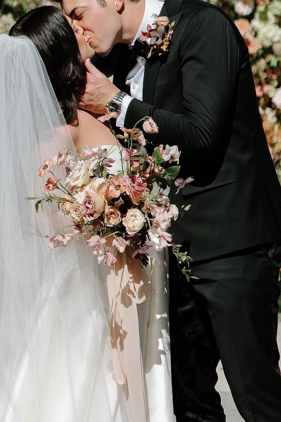 Wedding kiss portrait of bride and groom kissing, veil and rose bouquet in hand, framed by a floral arch in a lush garden backdrop