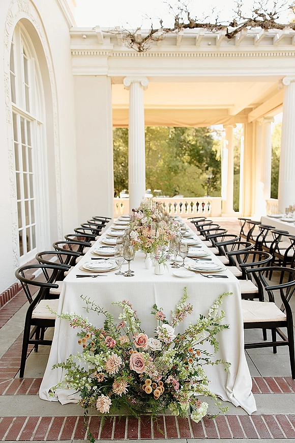 Reception tablescape with long banquet table decor, pastel floral centerpieces, bud vases, and black chairs on a covered veranda with columns