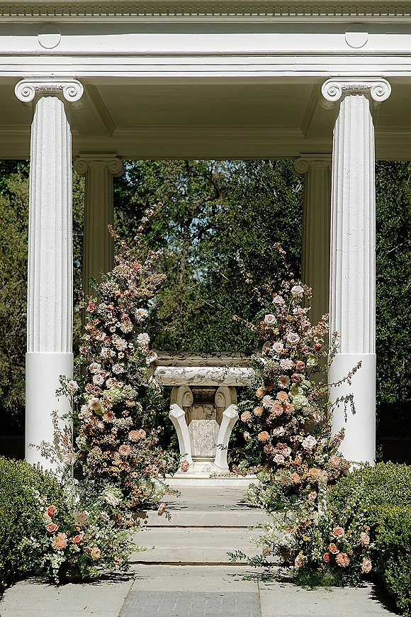 Ceremony altar decor with asymmetrical wedding arch flowers in roses, dahlias, and greenery on a stone pedestal before a white portico